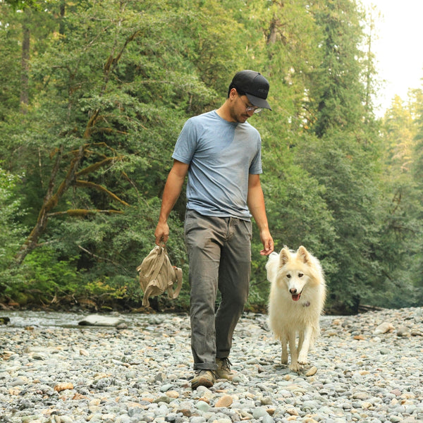 man in baseball cap walking white dog along rocky forest river
